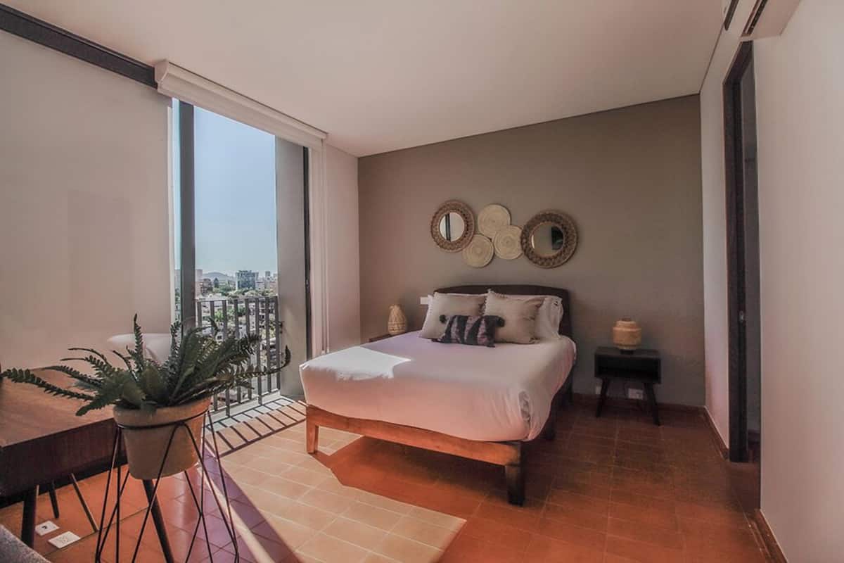 Un dormitorio con cama, una mesita con una planta, un ventanal con vistas y espejos decorativos en la pared.