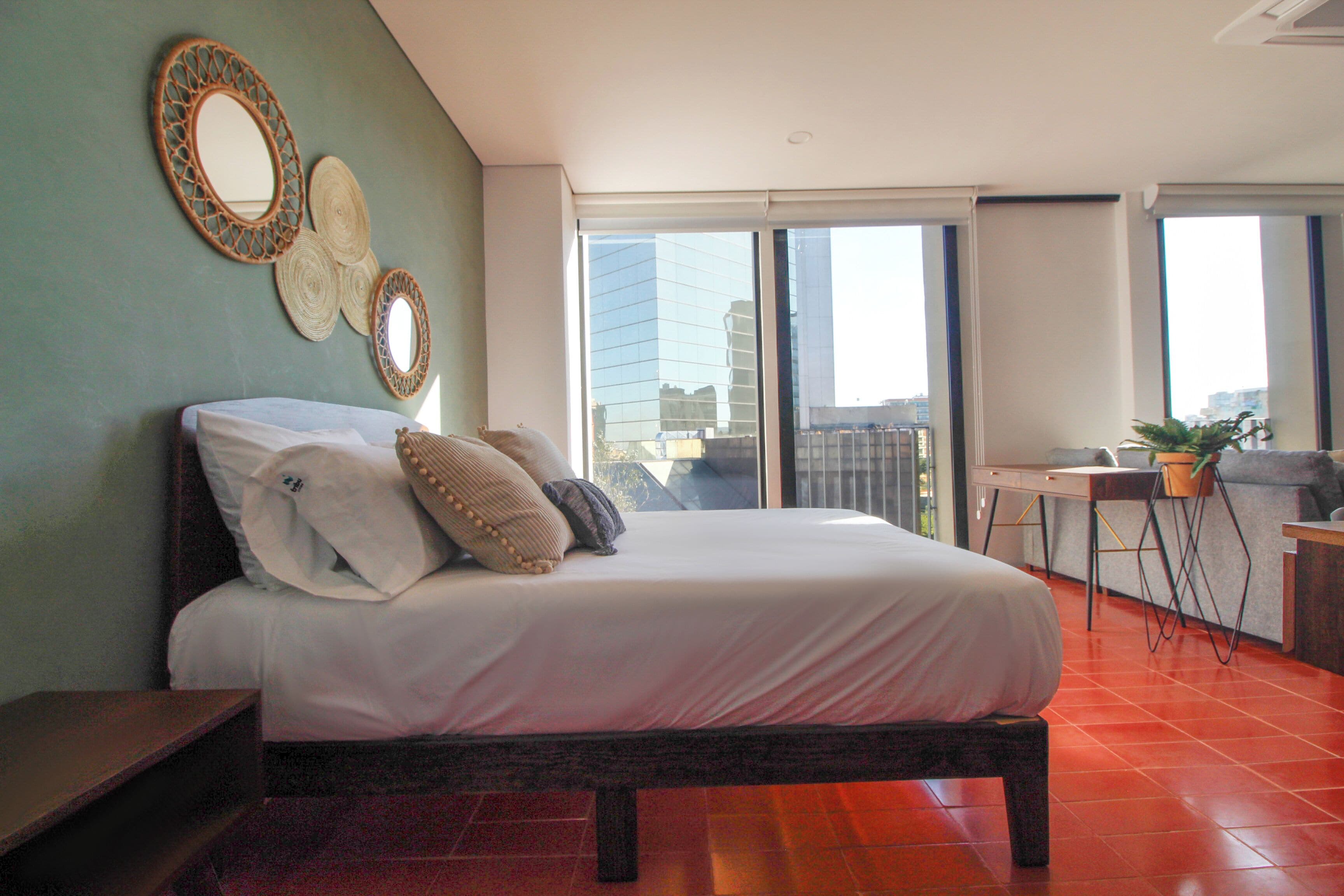 A modern bedroom with a large bed, a desk, and a view of the city through large windows.