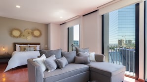 A modern hotel room with a grey sectional sofa, a large bed with white linens, and a view of a cityscape through large windows. - Tribu Chapultepec 67 (Guadalajara)