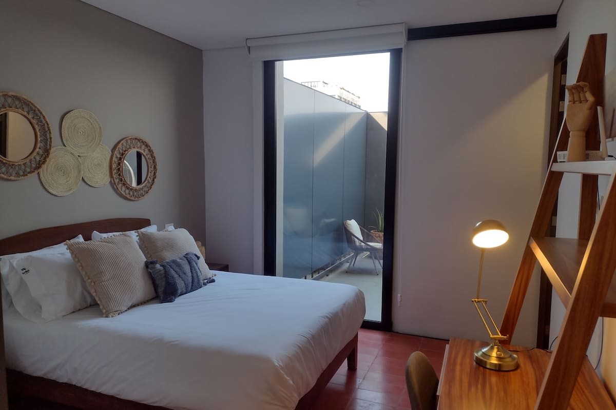 Un dormitorio con cama, mesita de noche, lámpara y vistas al exterior.