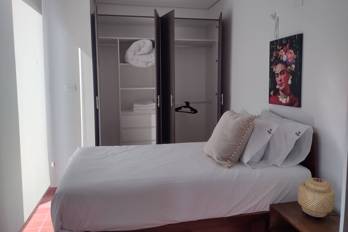 Un dormitorio con cama, armario, perchero, cojín decorativo, una obra de arte en la pared y una lámpara de fibras.