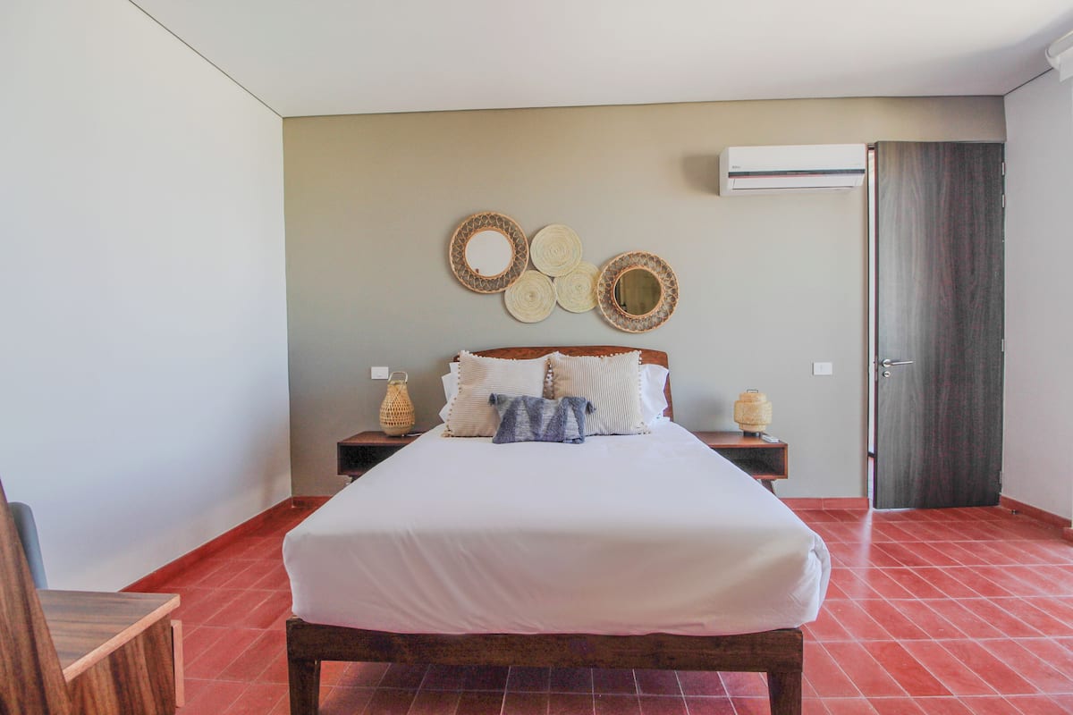 Un dormitorio ordenado con una cama grande, una mesita de noche de madera, un aire acondicionado instalado en la pared y una puerta.
