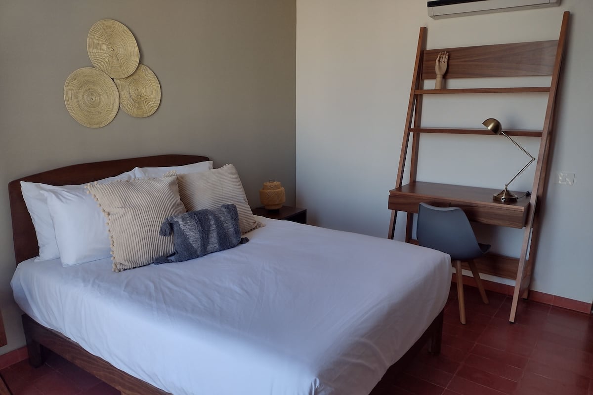 Una cama bien hecha con sábanas blancas y varios almohadones, un escritorio de madera con una silla, y una estantería en la pared.