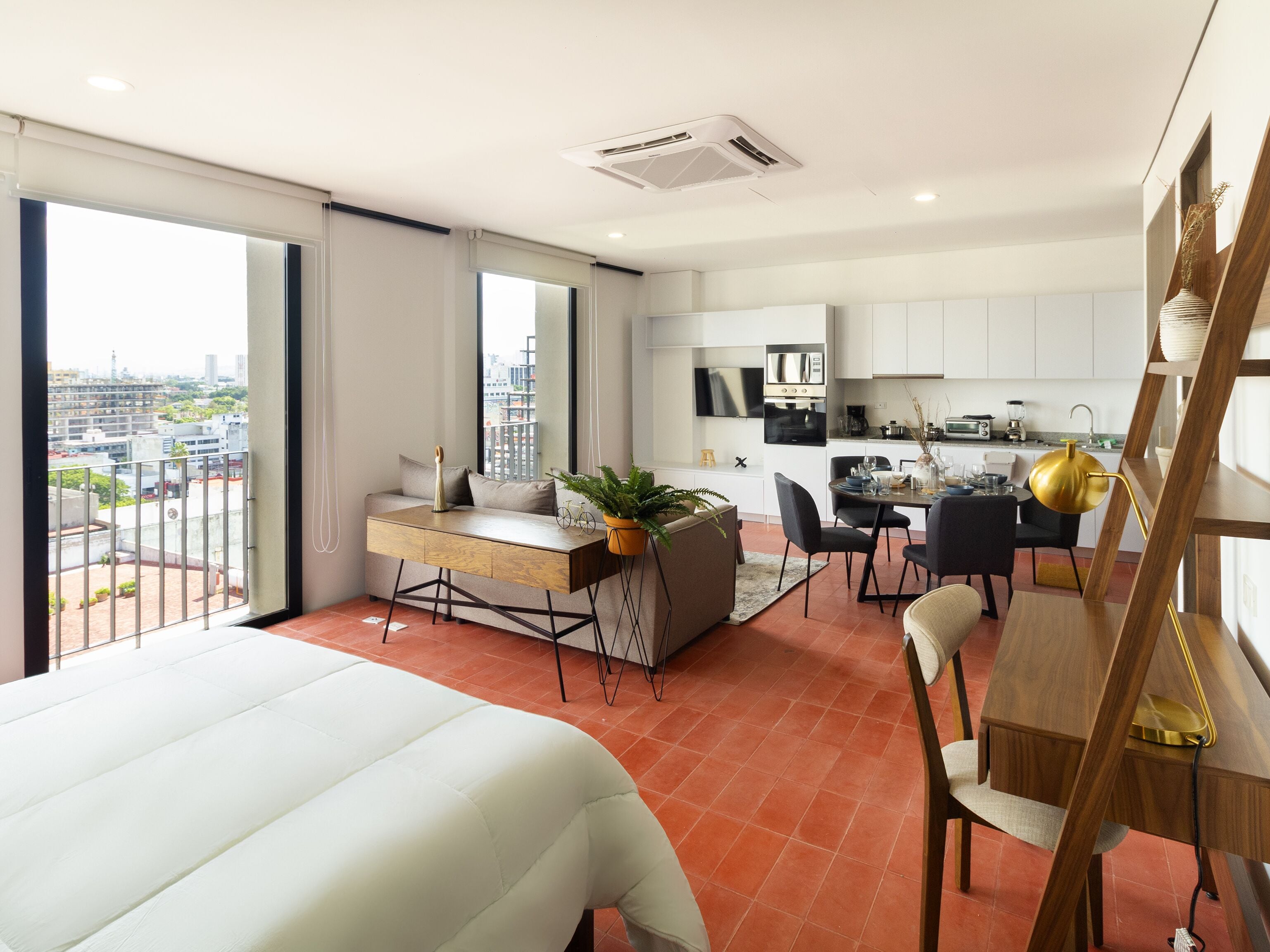 A modern hotel room with a large bed, a desk with a view, a dining area, and a kitchen.