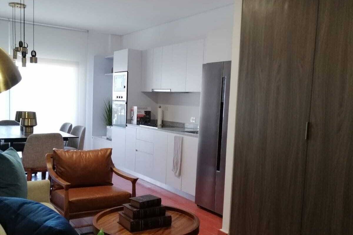 Una sala de estar moderna con un sofá, dos mesas auxiliares y una zona de comedor con una mesa y sillas. Se observa una cocina al fondo con armarios blancos y un refrigerador.