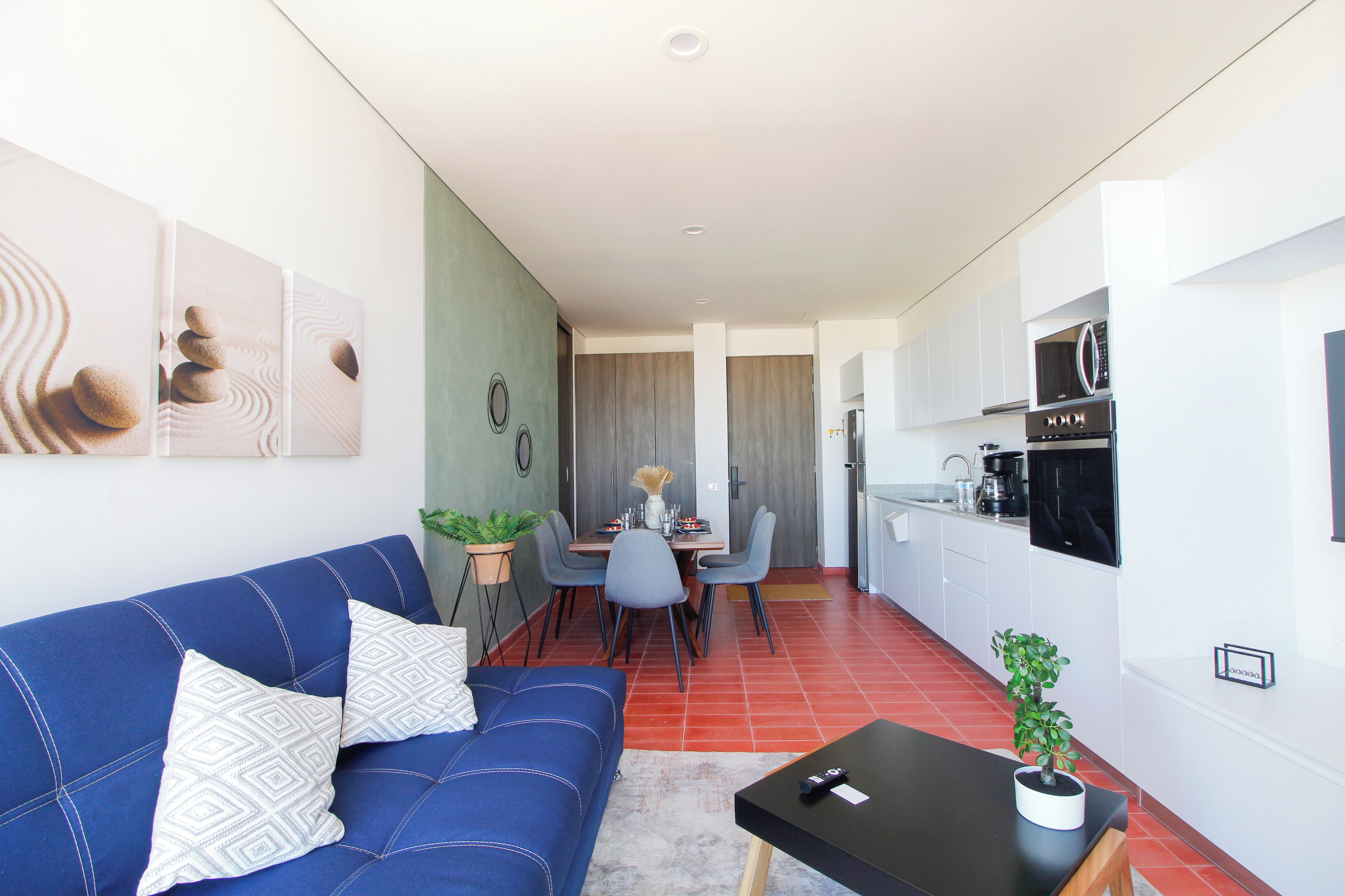 Un salon moderne avec un canapé bleu, une table à manger et des chaises. On y trouve également un coin cuisine avec des meubles blancs et un four à micro-ondes.