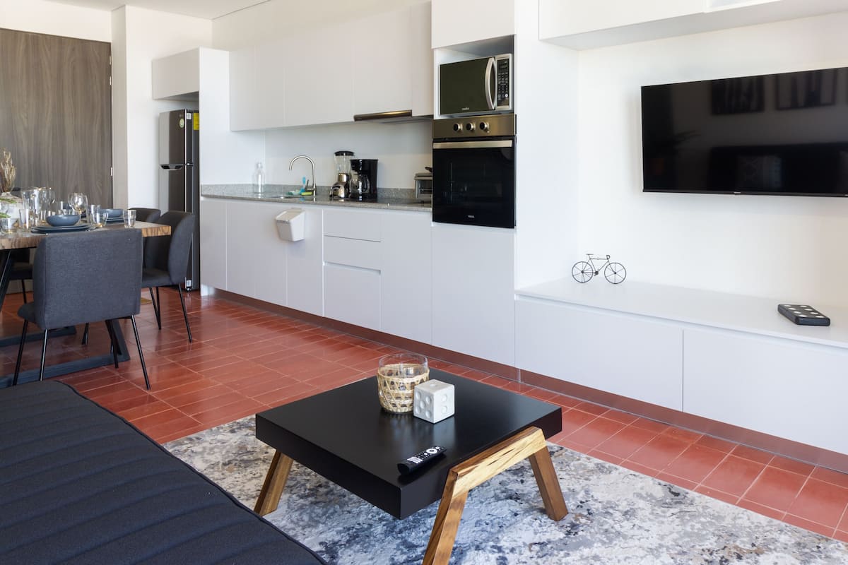 Una cocina moderna con armarios blancos, un microondas empotrado y una estufa. Hay una zona de comedor con una mesa y sillas, y una mesa de centro negra con un control remoto.