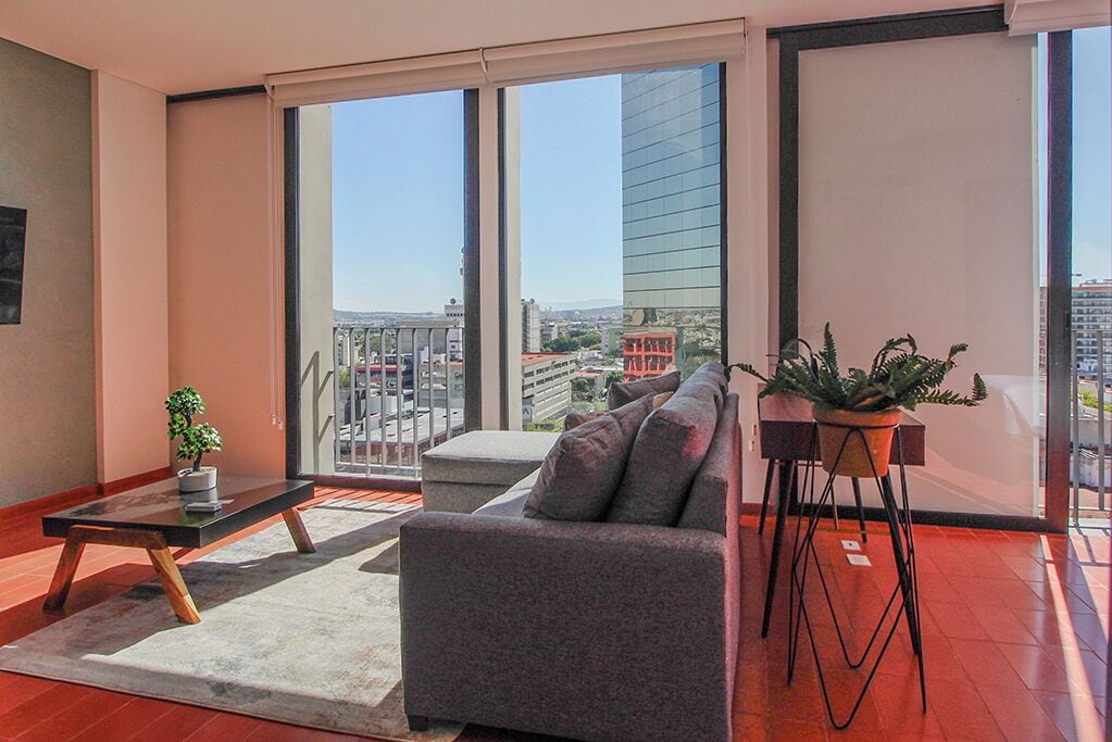 A modern living room with a grey sofa, a small table with a plant, and a large window offering a city view.
