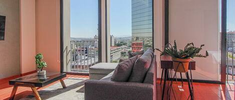 A modern living room with a grey sofa, a small table with a plant, and a large window offering a city view.