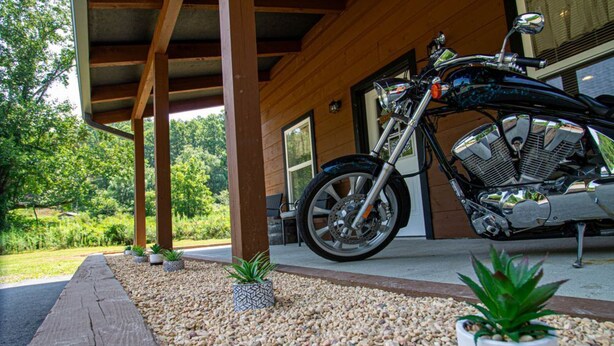Una motocicletta parcheggiata sotto un pergolato in legno, con un vialetto sterrato e piante in vaso.