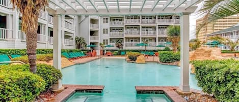 Un área de piscina con una sección cubierta, sillas de descanso y un edificio con balcones al fondo.