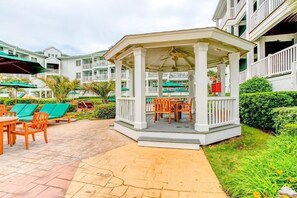 A covered outdoor seating area with a table and chairs, surrounded by greenery and a poolside lounge. - Virginia Beach | Couples Ocean Coast Retreat | Pool + Hot Tub (Virginia Beach)