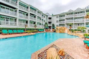 Piscina rodeada por um edifício com vários andares, varandas, espreguiçadeiras e um jardim com pedras e plantas.