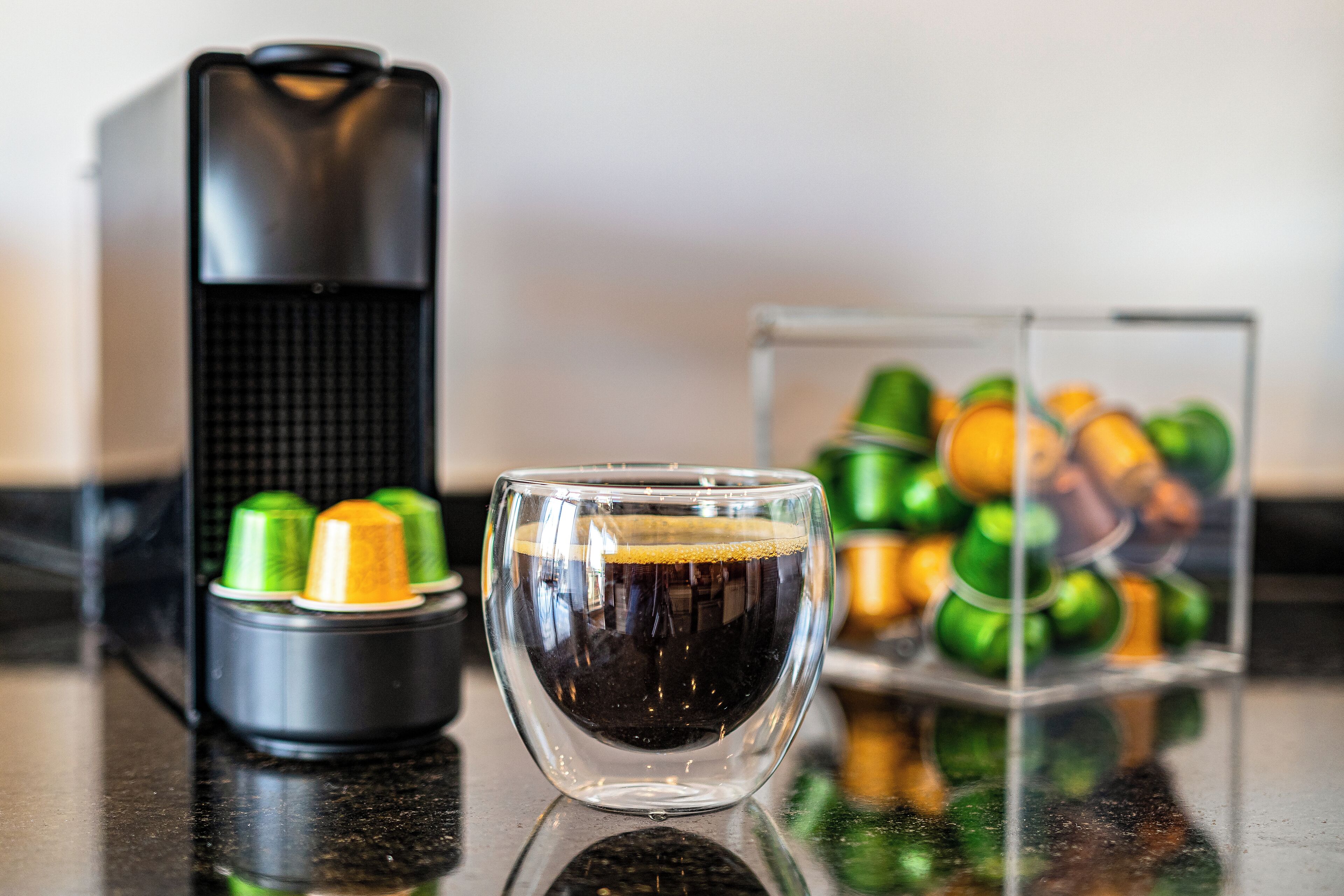 Máquina de café, chávena com café e um recipiente com cápsulas de café numa bancada.