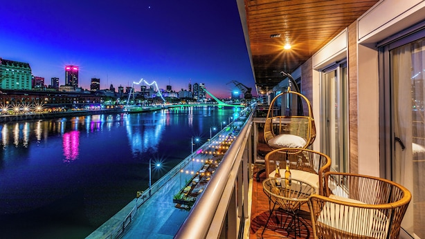 A balcony with wicker chairs and a table, overlooking a river with city lights and a bridge. - 2BR Family Apt Puente de la Mujer View (Buenos Aires)
