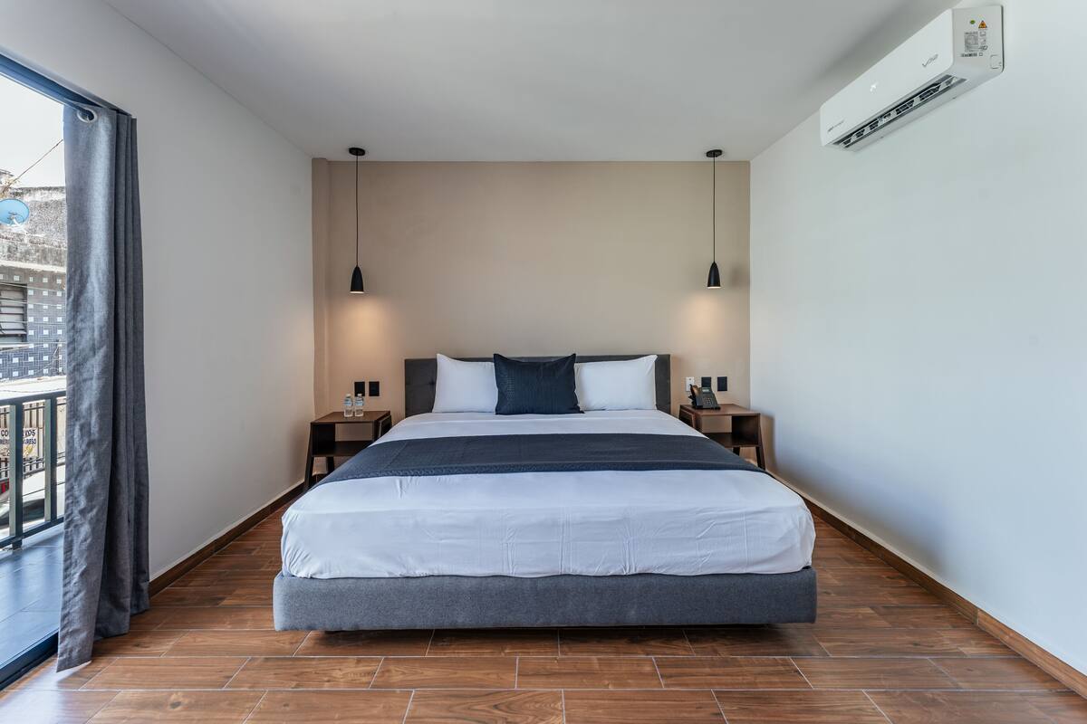 A modern hotel room with a large bed, two bedside tables, a nightstand, a window with curtains, and an air conditioning unit on the wall.