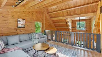 A cozy cabin interior with a grey sofa, two round wooden tables, and a balcony with a railing.