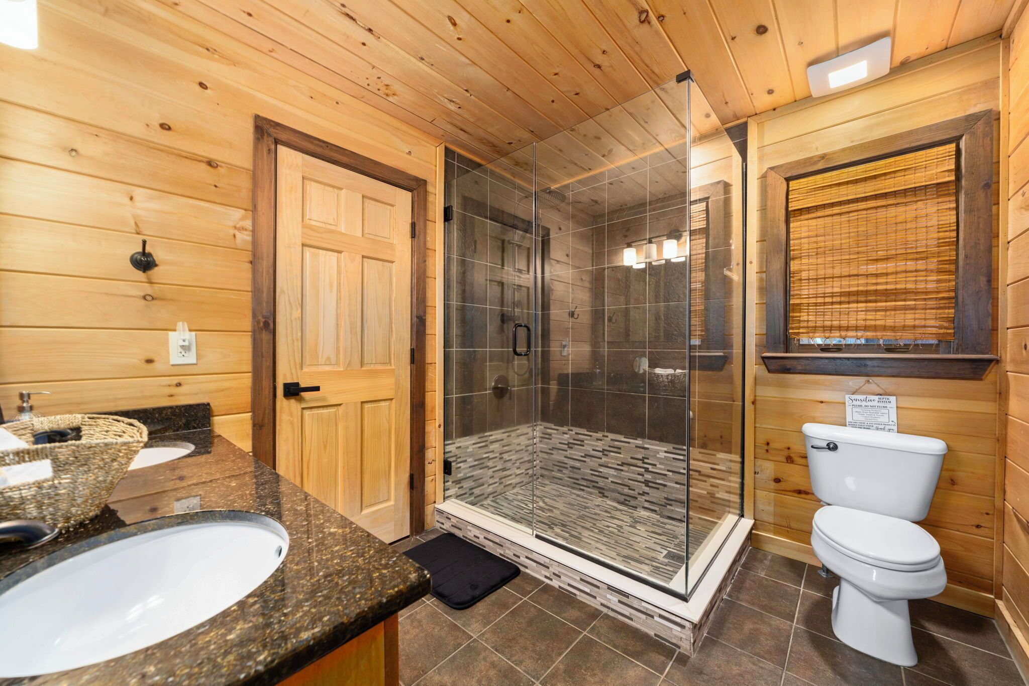 Une salle de bain rustique, avec des murs en bois, une douche en verre, deux lavabos et des toilettes.