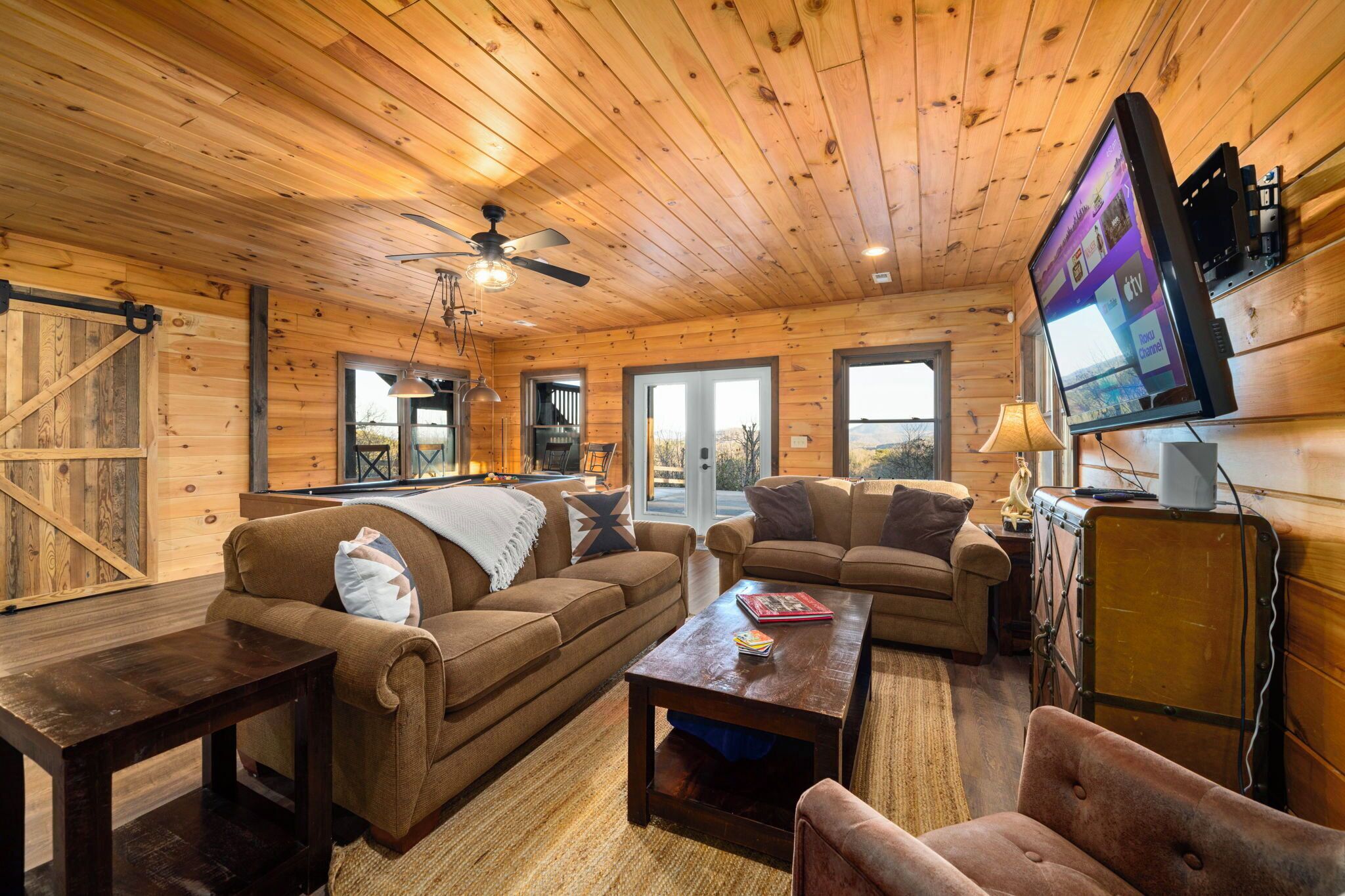 A cozy cabin interior with wooden walls, a ceiling fan, a flat-screen TV mounted on the wall, and a large window offering a view of the outdoors.
