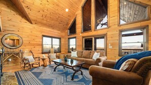 A cozy cabin interior with wooden walls, a large window, a sofa, a rocking chair, and a coffee table. - 10% Off Member Deal•OMG Views•10 Min to Downtown•Private•Hot Tub•Deck•Firepit (Blue Ridge)