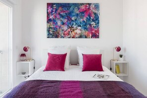 A bedroom with a white bed, two pink pillows, a purple blanket, and a floral painting above the bed.