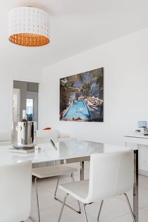 A modern dining area with a glass-top table, white chairs, and a large painting of a pool scene. - Stroll to Beach from Sea-View Penthouse with AC (Antibes)