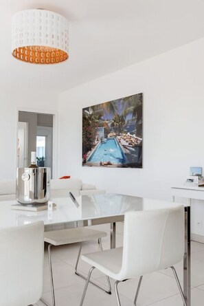 A modern dining area with a glass table, white chairs, a large painting of a pool, and a pendant light.