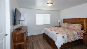 Une chambre à coucher avec un lit en bois, un téléviseur fixé au mur, une fenêtre avec des stores et une commode en bois.
