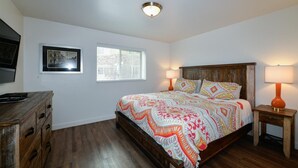 Quarto com cama de madeira, duas luminárias de cabeceira, cômoda, quadro emoldurado e janela.