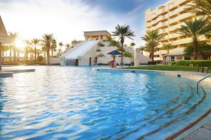 A swimming pool with a waterfall feature, surrounded by palm trees and a building with balconies.