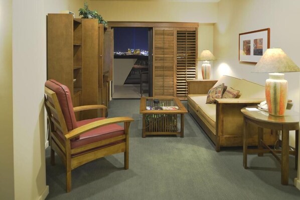 Un salón acogedor con un sofá de madera, un sillón rojo, una mesita de centro y una lámpara. Posee una puerta corredera que da a un balcón con vistas a la ciudad de noche.