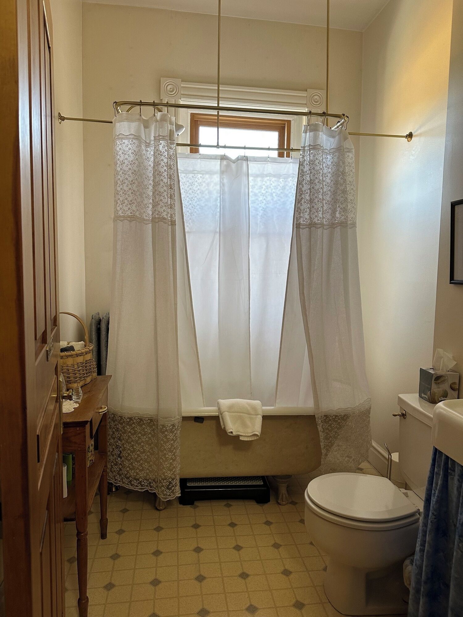 Une salle de bain avec des toilettes, une fenêtre avec des rideaux légers, un meuble-lavabo en bois et un sol carrelé.