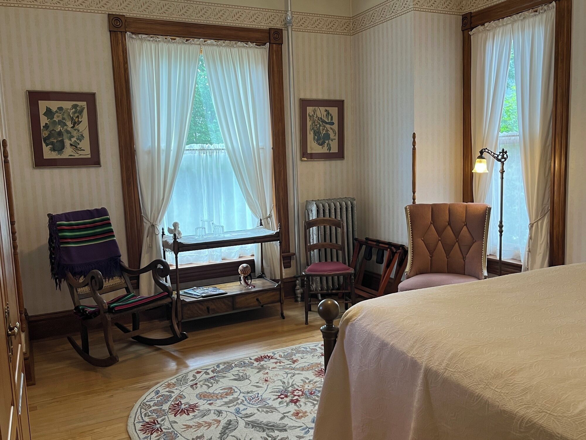 A bedroom with a bed, rocking chair, chair with a cushion, a small table, and a floor lamp.