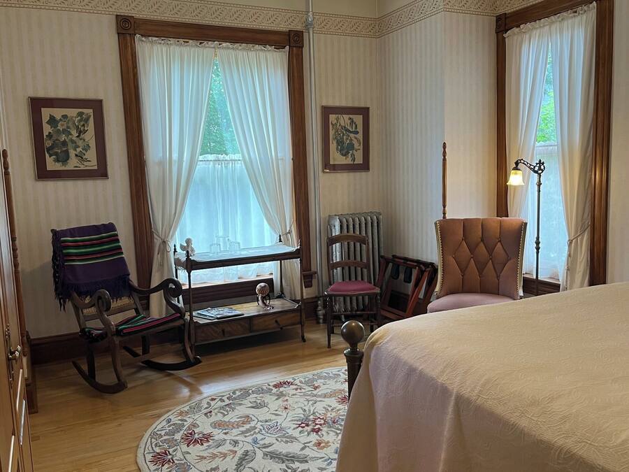 A bedroom with a bed, rocking chair, chair with a cushion, a small table, and a floor lamp.