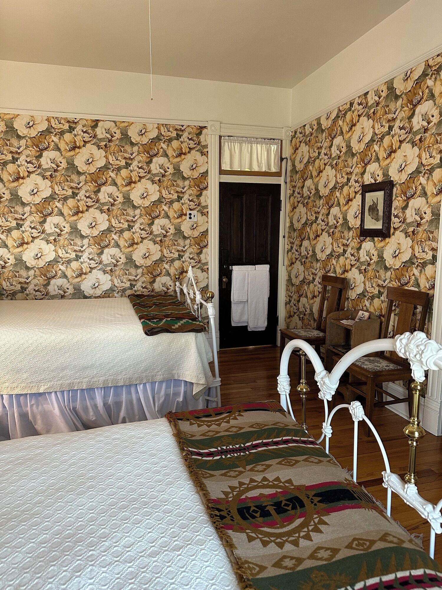 A bedroom with two beds, floral wallpaper, a wooden floor, and a door leading to a bathroom.