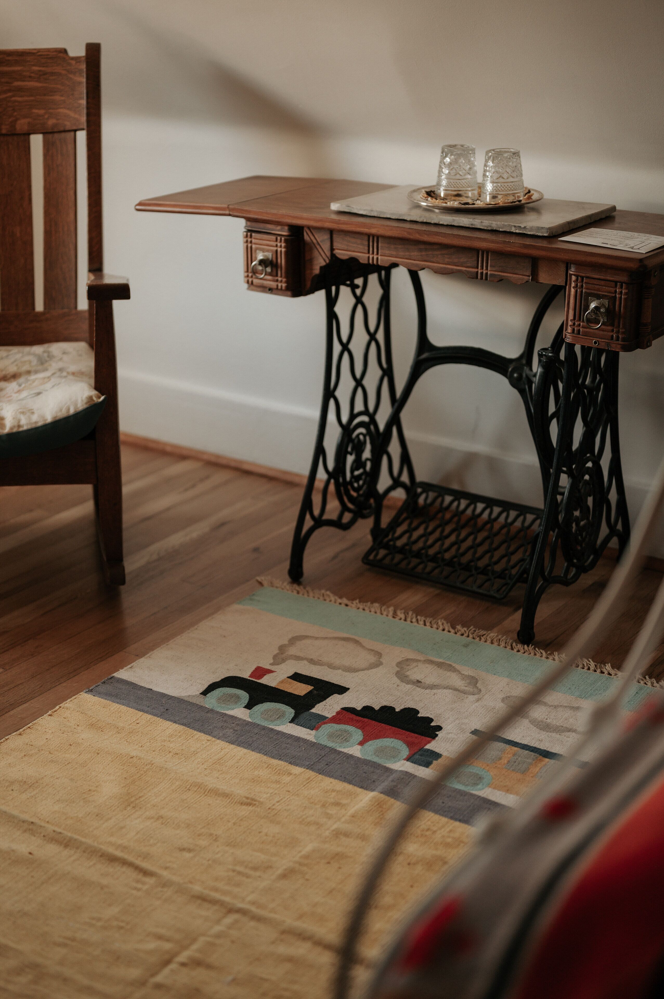 Una silla de madera con cojín, una mesa de máquina de coser antigua con dos vasos de vidrio, y una alfombra estampada sobre piso de madera.