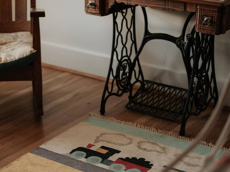 Una silla de madera con cojín, una mesa de máquina de coser antigua con dos vasos de vidrio, y una alfombra estampada sobre piso de madera.