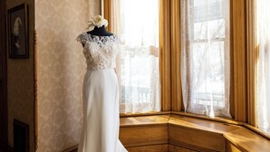 Une robe de mariée est exposée sur un mannequin dans une pièce dotée d’une grande fenêtre, d’un plancher en bois et d’un tableau accroché au mur.