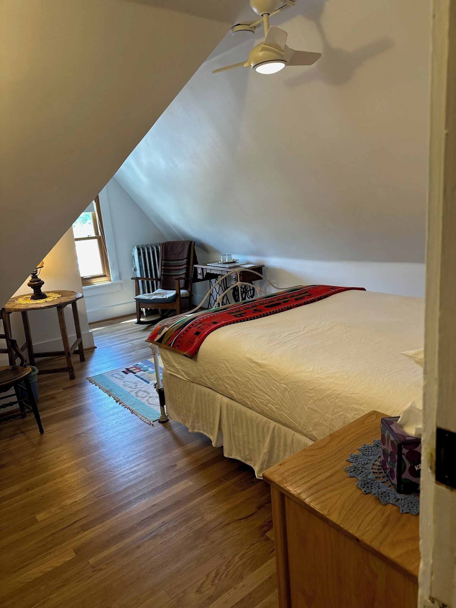A bedroom with a sloped ceiling, a bed with a red blanket, a wooden table, a chair, and a small table with a lamp.