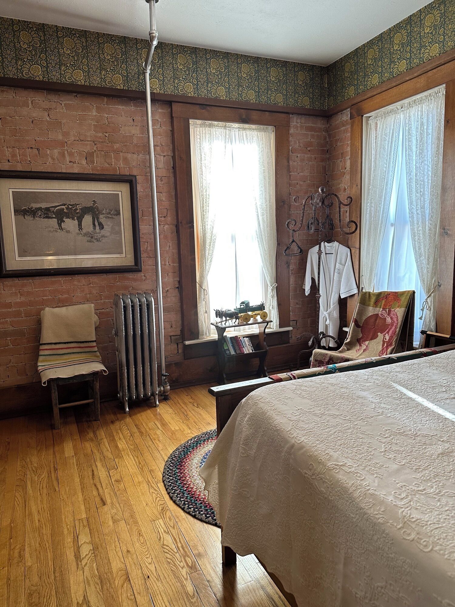 Une pièce avec un mur en briques, une photo historique encadrée, un radiateur, un plancher en bois, un lit recouvert d’une couvre-lit à motifs et une fenêtre ornée de rideaux légers.