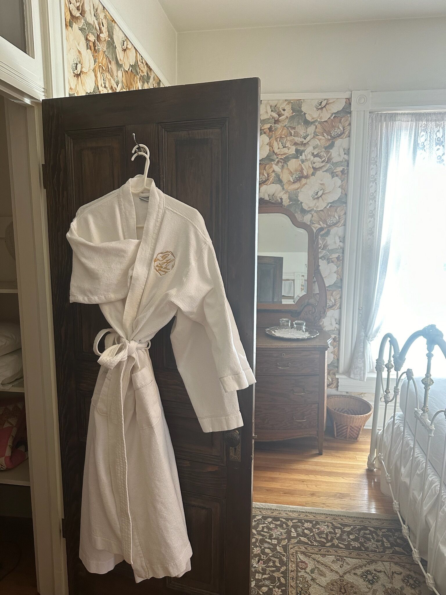 A white robe hanging on a door, a wooden dresser, a mirror, a bed with white bedding, and a floral wallpaper.