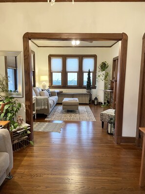 A spacious living room with a wooden floor, a sofa, a coffee table, and a fireplace. There are large windows with blinds, a potted plant, and a mirror. - A charming craftsman located in the perfect American small town (Saint Lucas)