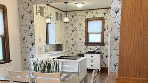 A dining area with a wooden table and chairs, a chandelier, and a kitchen in the background with floral wallpaper. - A charming craftsman located in the perfect American small town (Saint Lucas)