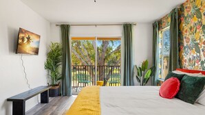 Quarto com cama, um banco, televisão e varanda com cadeiras e plantas.