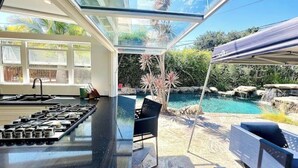 Et køkken med en stor ø, et spiseområde med stole, og udsigt til en baghave med pool og vandfald.