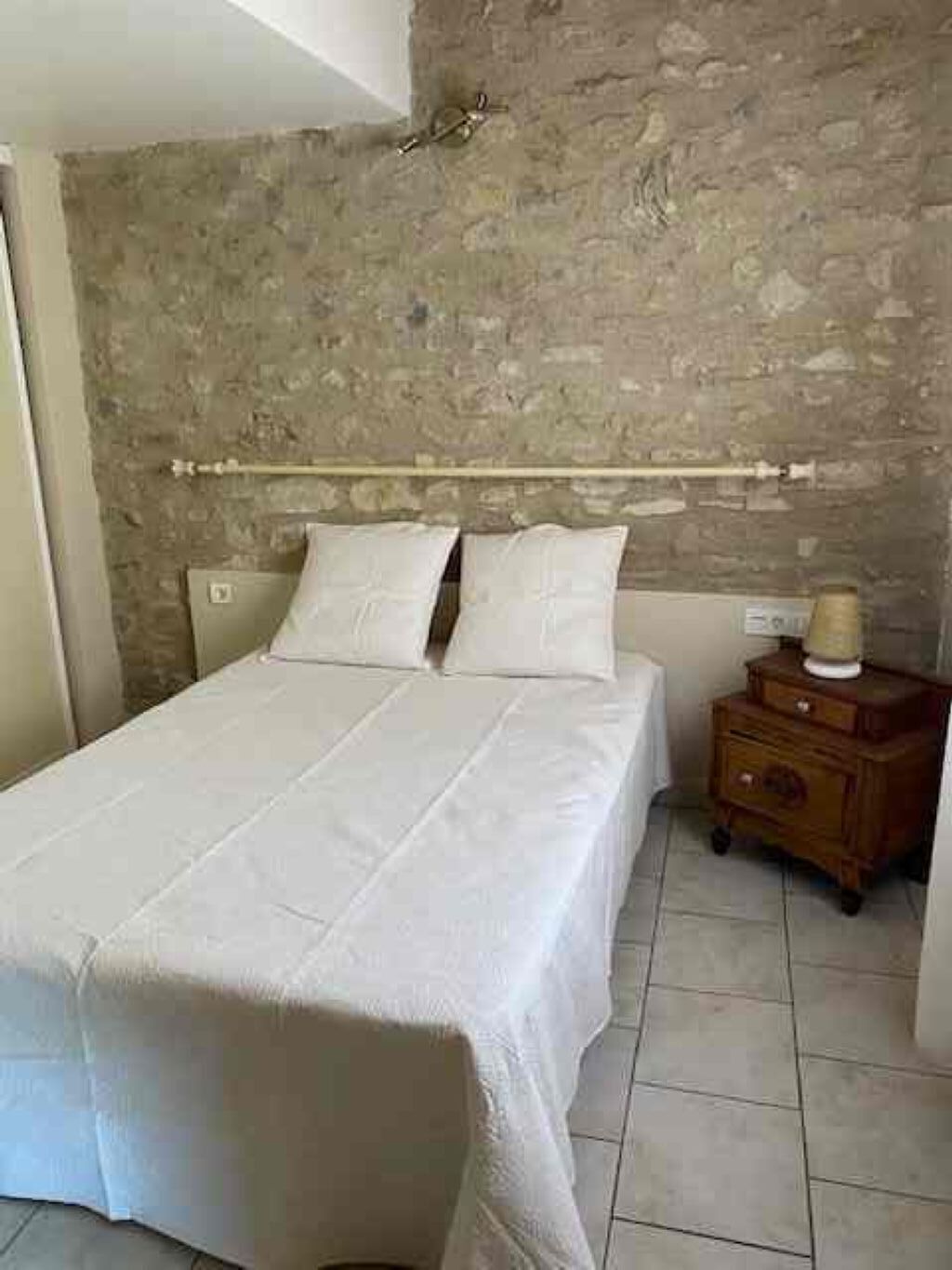 A bedroom with a stone wall, a bed with white bedding, a wooden nightstand, and a lamp.
