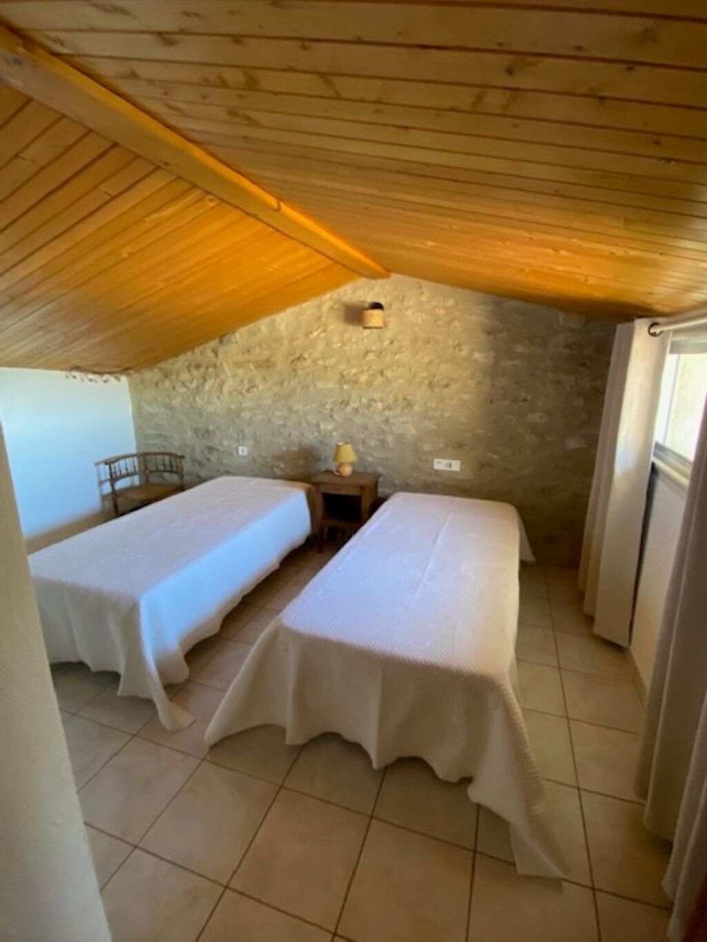 A small, cozy room with two beds, a wooden ceiling, a stone wall, and a small table with a lamp.