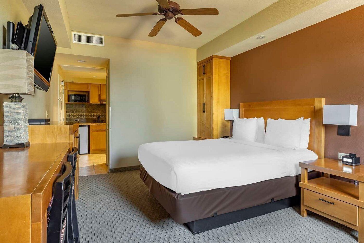 Una habitación de hotel con cama, mesitas de noche, un escritorio, un ventilador de techo y vistas a la cocina.