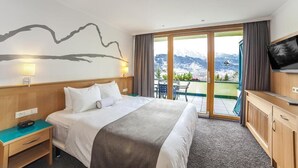 A hotel room with a bed, a desk, a TV, and a balcony with a mountain view. - Schladming | Couples Ski Stop | Pool + Spa (Schladming)
