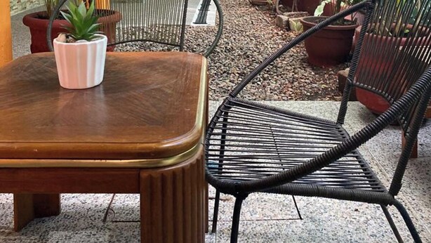 Un espacio al aire libre techado con una mesa de madera y sillas de ratán negro, rodeado de plantas en macetas y una pared verde.